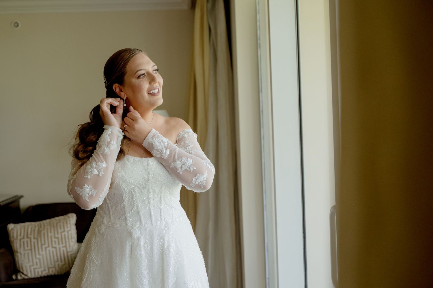 A bride in a white lace long-sleeve wedding dress puts on earrings while standing by a window.