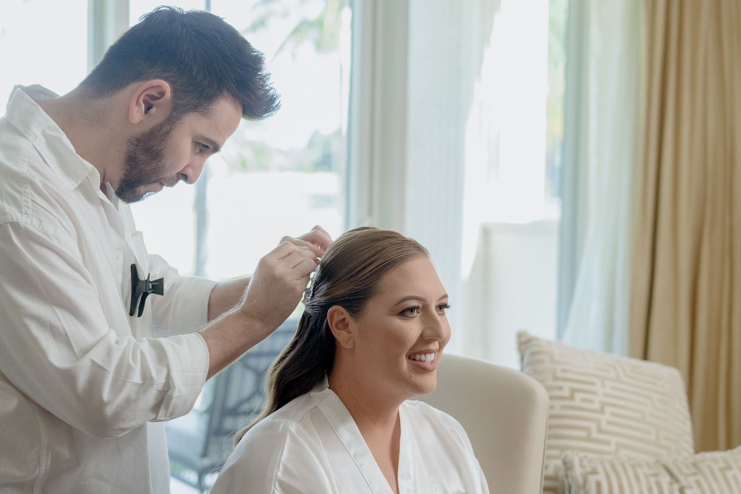 A hairstylist works on styling a client's hair in a bright, sunlit room while wearing a white robe.