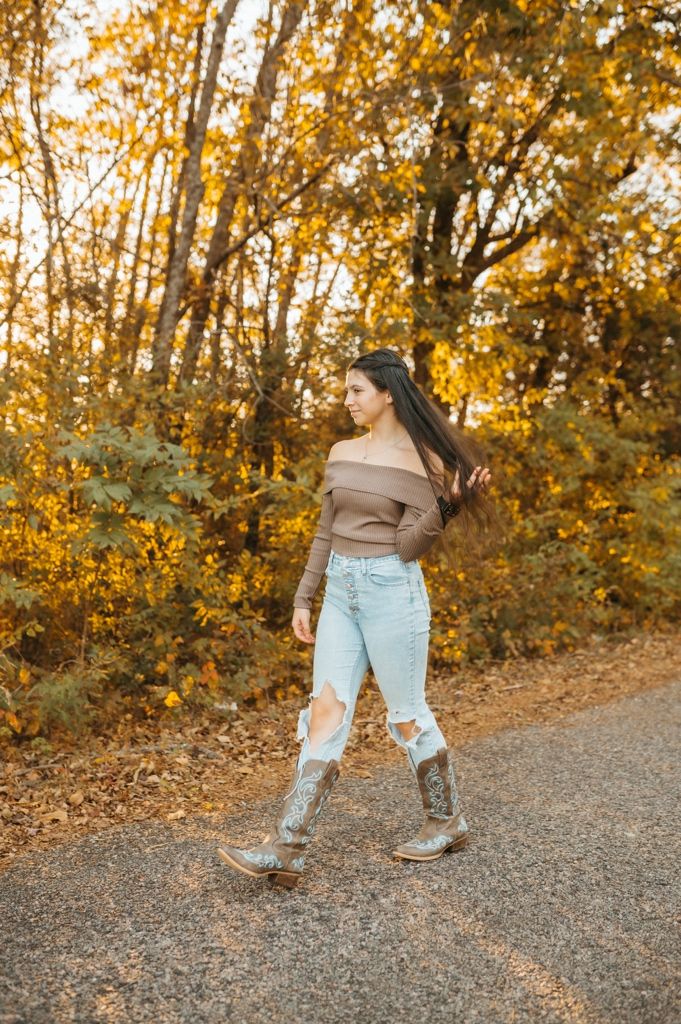 Person in brown top and ripped jeans dancing on gravel path surrounded by fall foliage.