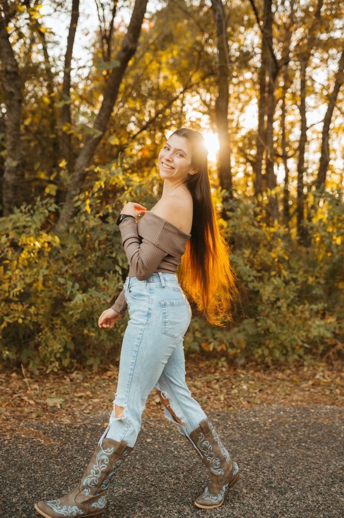 Person in brown sweater and light jeans poses playfully on gravel path during golden autumn sunset.