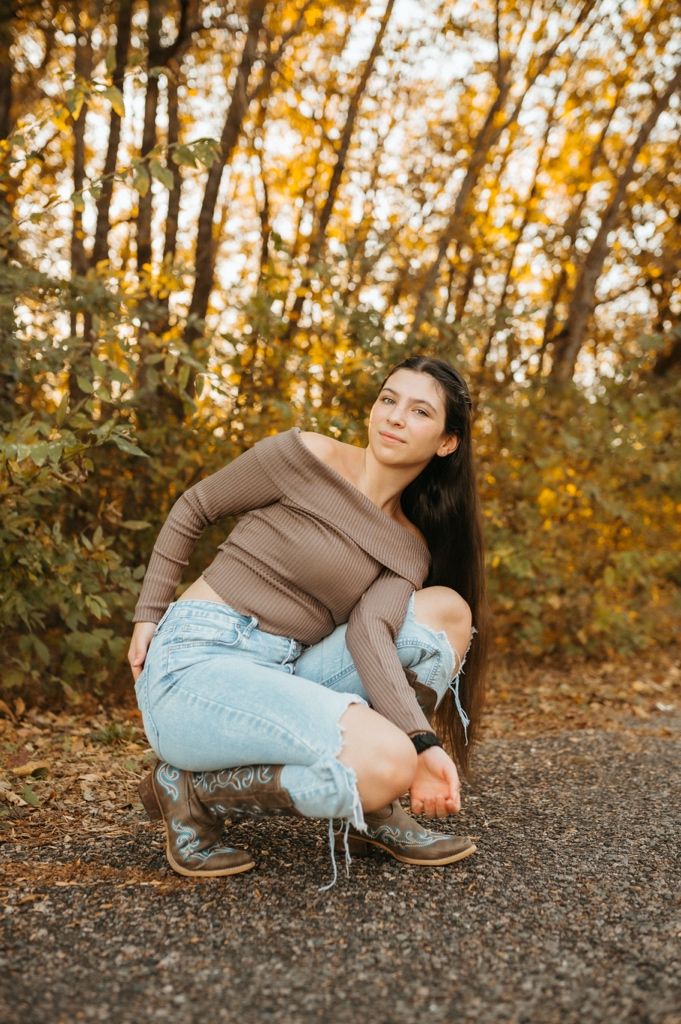 Person in brown top and distressed jeans sits casually on gravel path during autumn sunset.