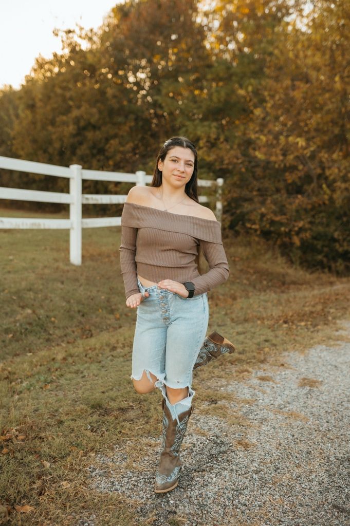Casual autumn outfit with brown off-shoulder top, distressed jeans and boots against white fence and fall foliage.