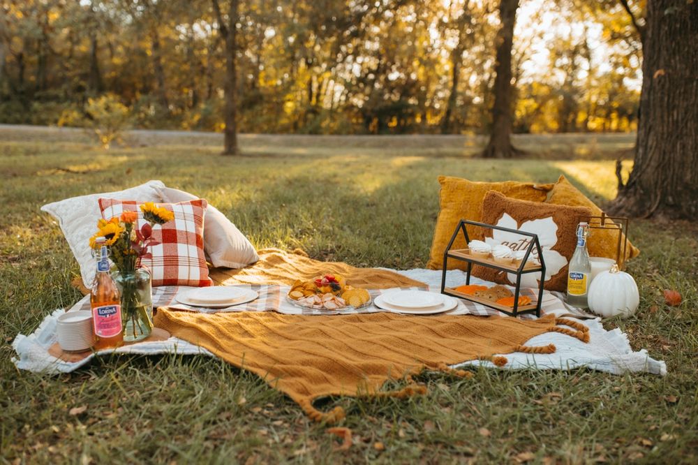 A cozy autumn picnic setup with blankets, pillows, and refreshments on grass during golden hour sunset.