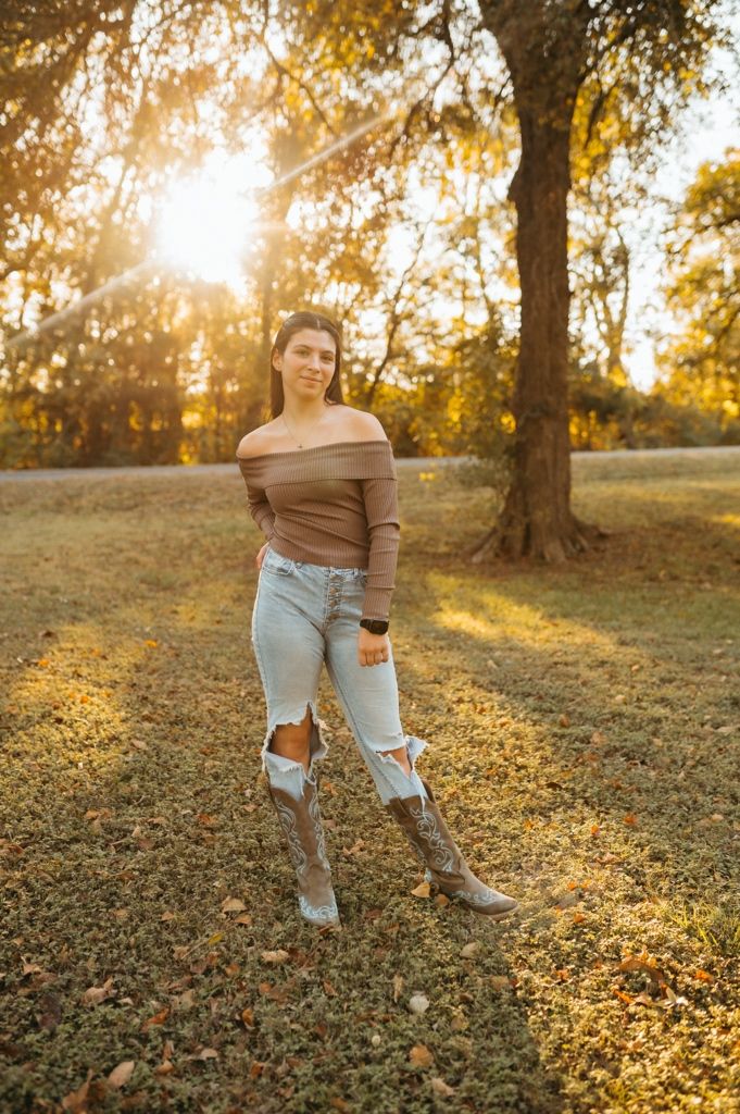 Person wearing ripped jeans and brown sweater stands in autumn field at golden hour with sunlight filtering through trees.