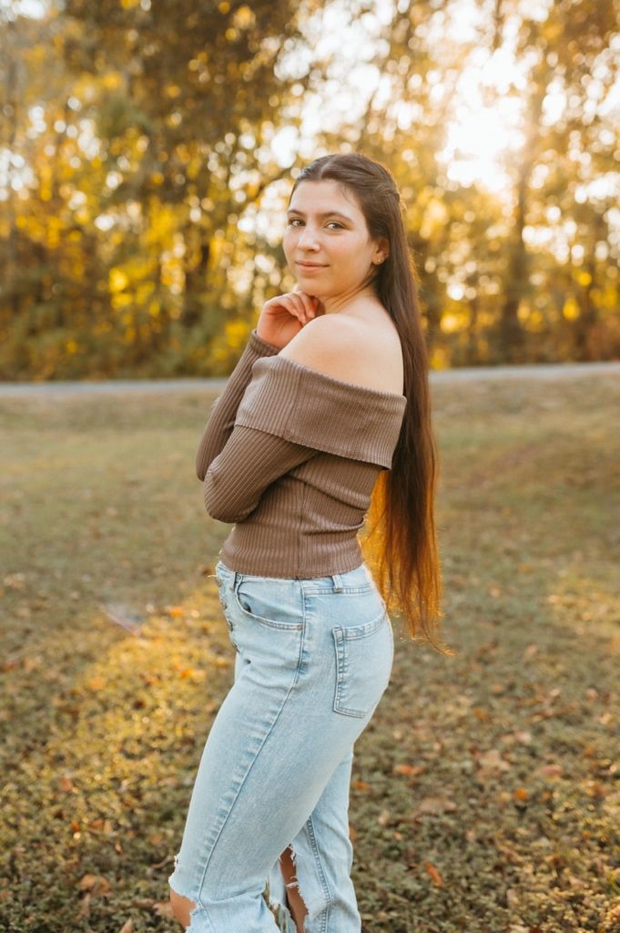 A person wearing a brown sweater and light jeans poses in an autumn field with golden sunlight filtering through trees.