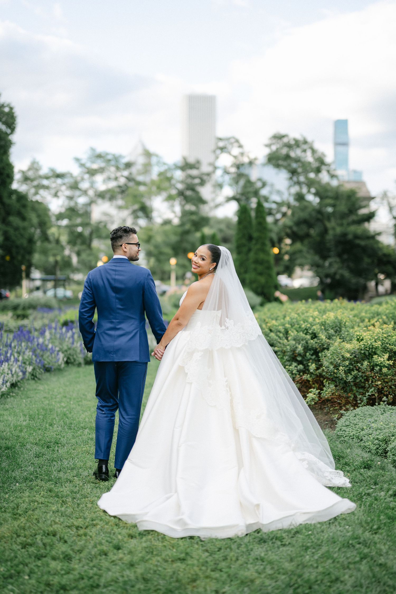 A couple in wedding attire stands on a manicured lawn with city buildings and trees in the background.