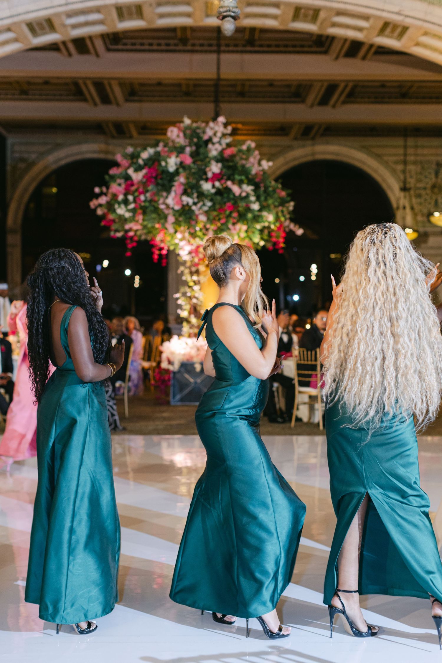 A sequence of bridesmaids in teal dresses dancing in a line at an elegant ballroom reception venue.