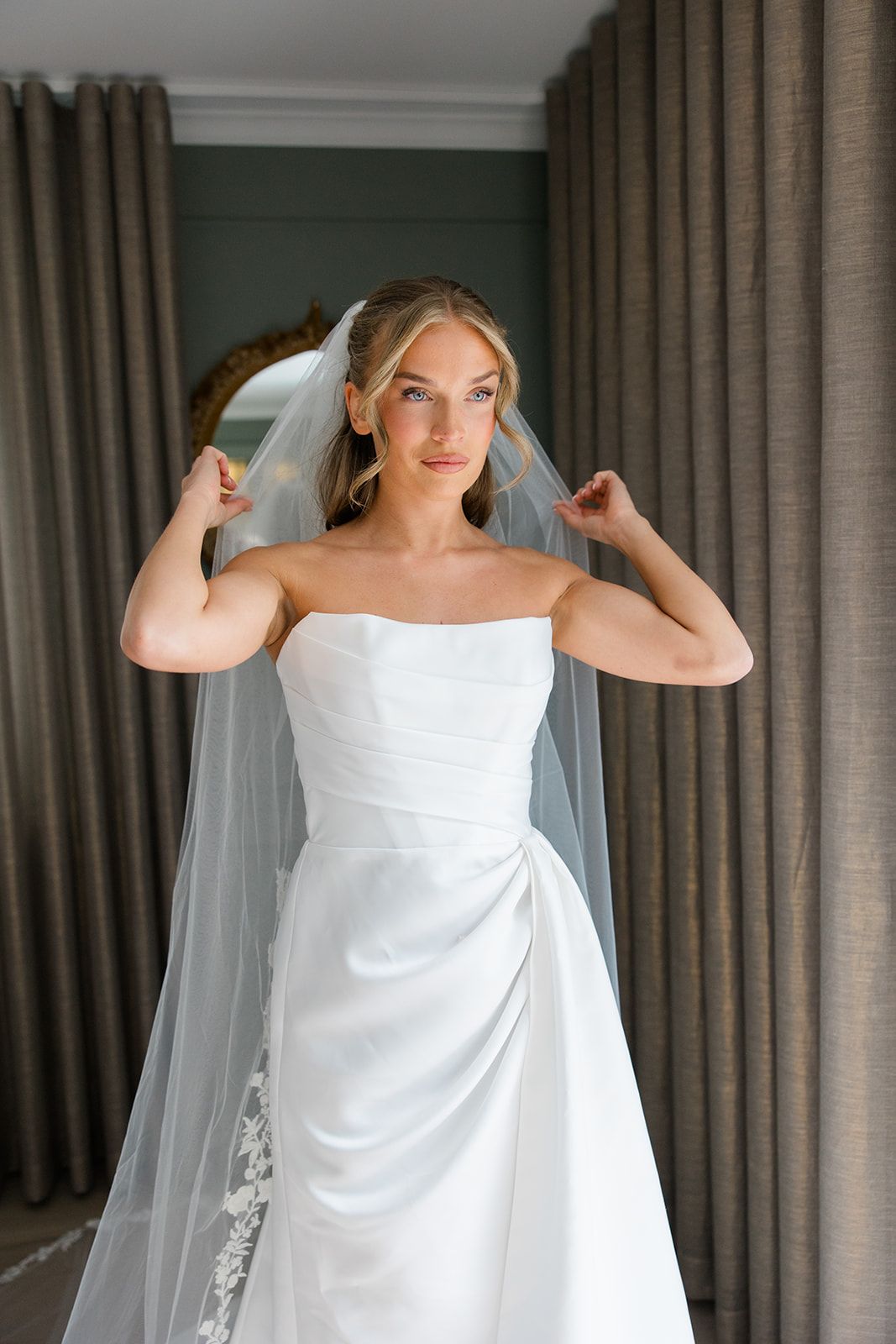 Bride in elegant white strapless wedding dress adjusts her flowing veil against a backdrop of gray curtains.