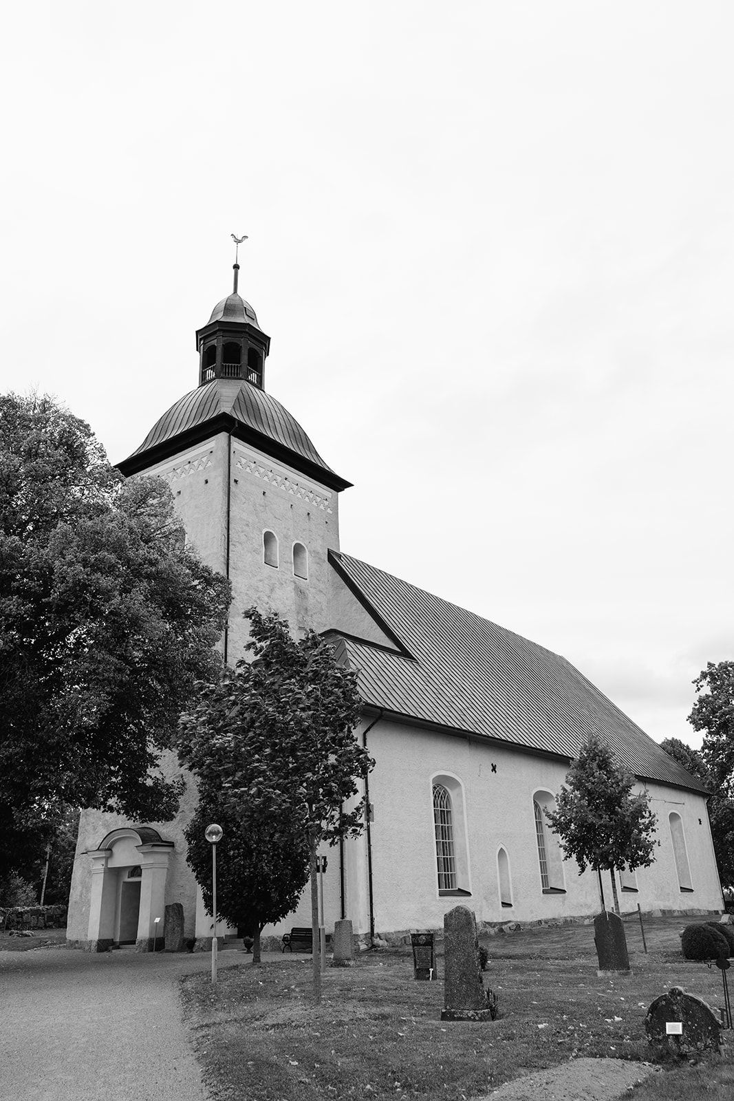 A black and white photograph of a historic white church with a tall spire and surrounding cemetery in a rural setting.
