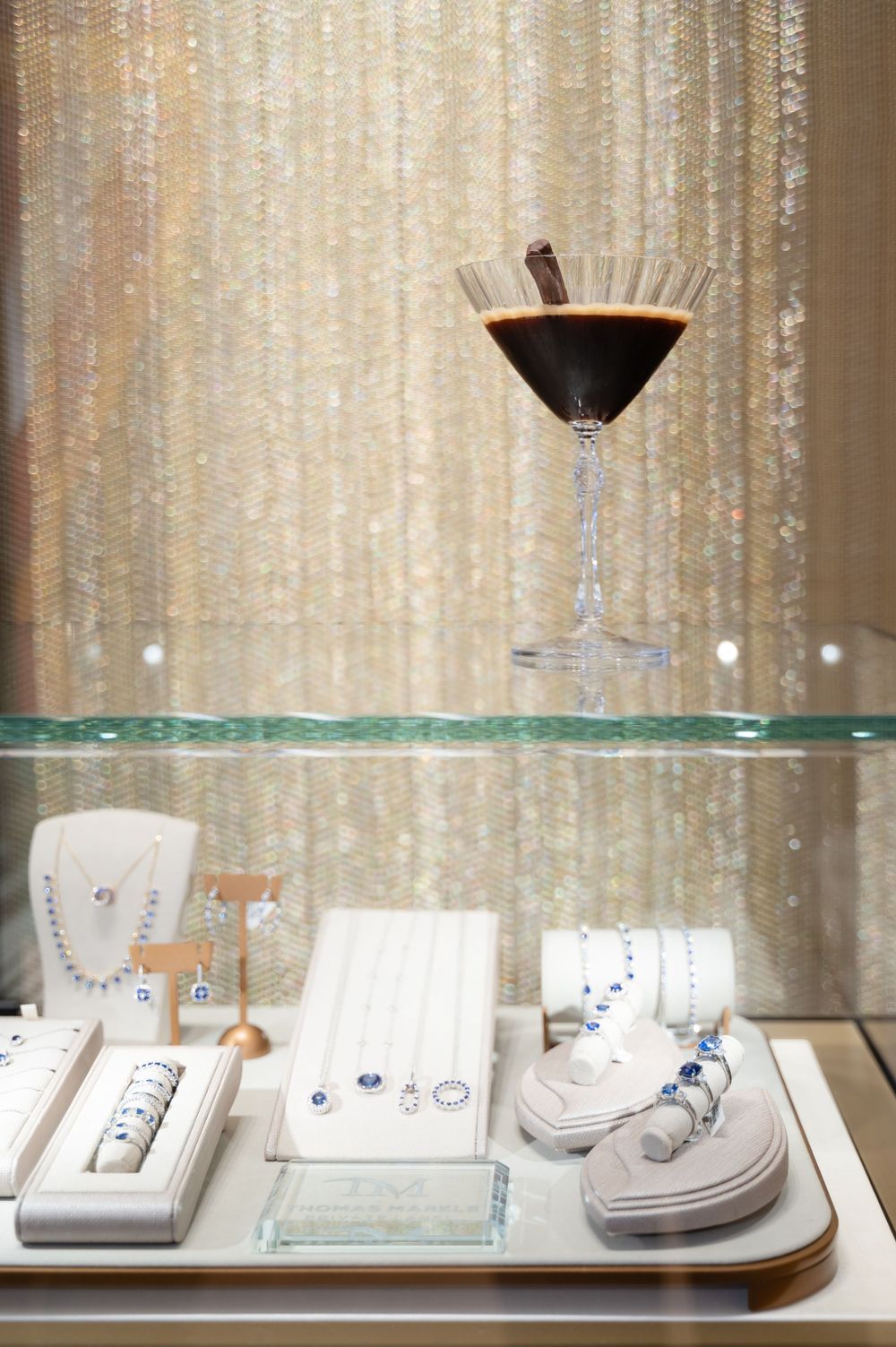 Elegant jewelry display with cocktail glass on glass shelf against shimmering gold curtain backdrop.