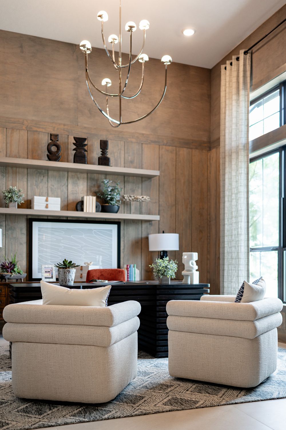 Modern living room with cream swivel chairs, contemporary chandelier, and wood-paneled walls featuring built-in shelving.