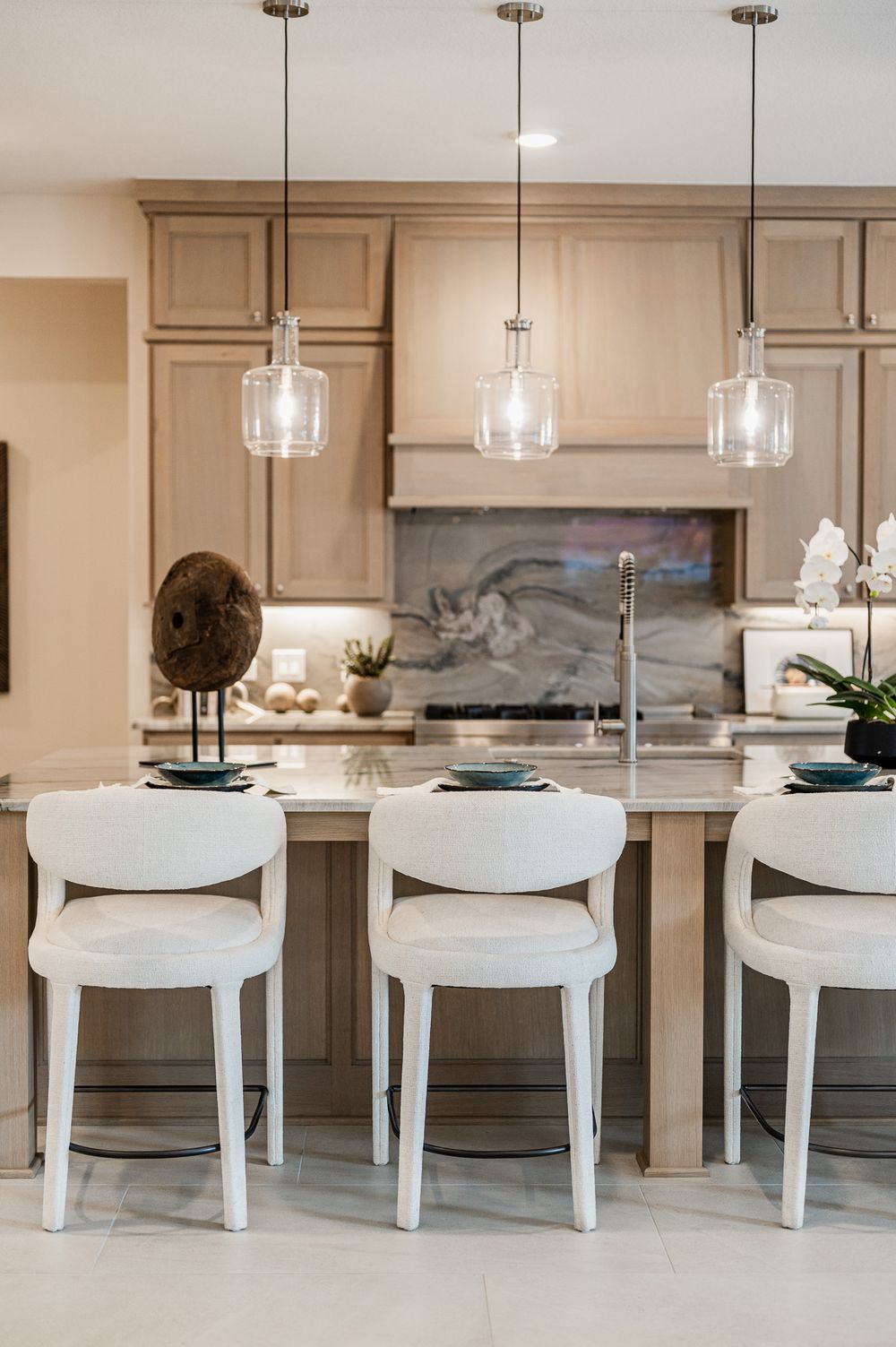 Modern kitchen island with six white chairs and glass pendant lights hanging above marble countertops.