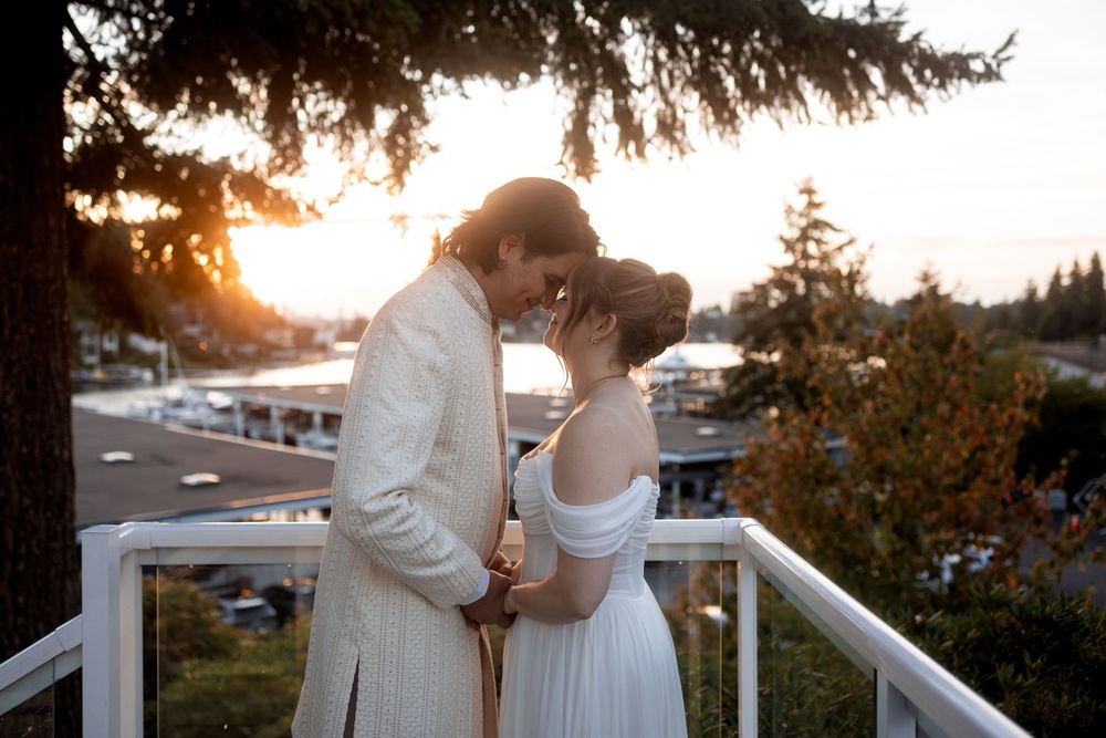 Newlyweds embrace on outdoor deck with waterfront and autumn trees in background.
