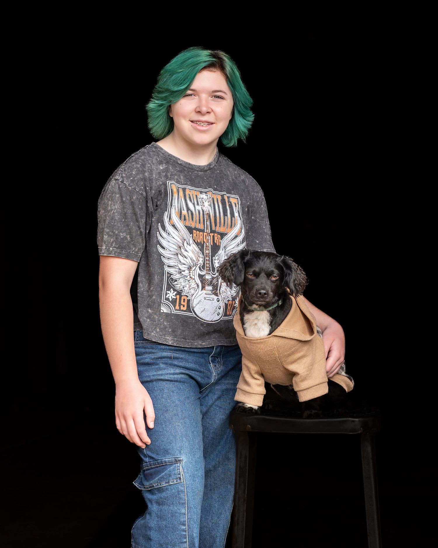 A person wearing a gray t-shirt and jeans poses with a small black dog wearing a brown outfit on a black chair.
