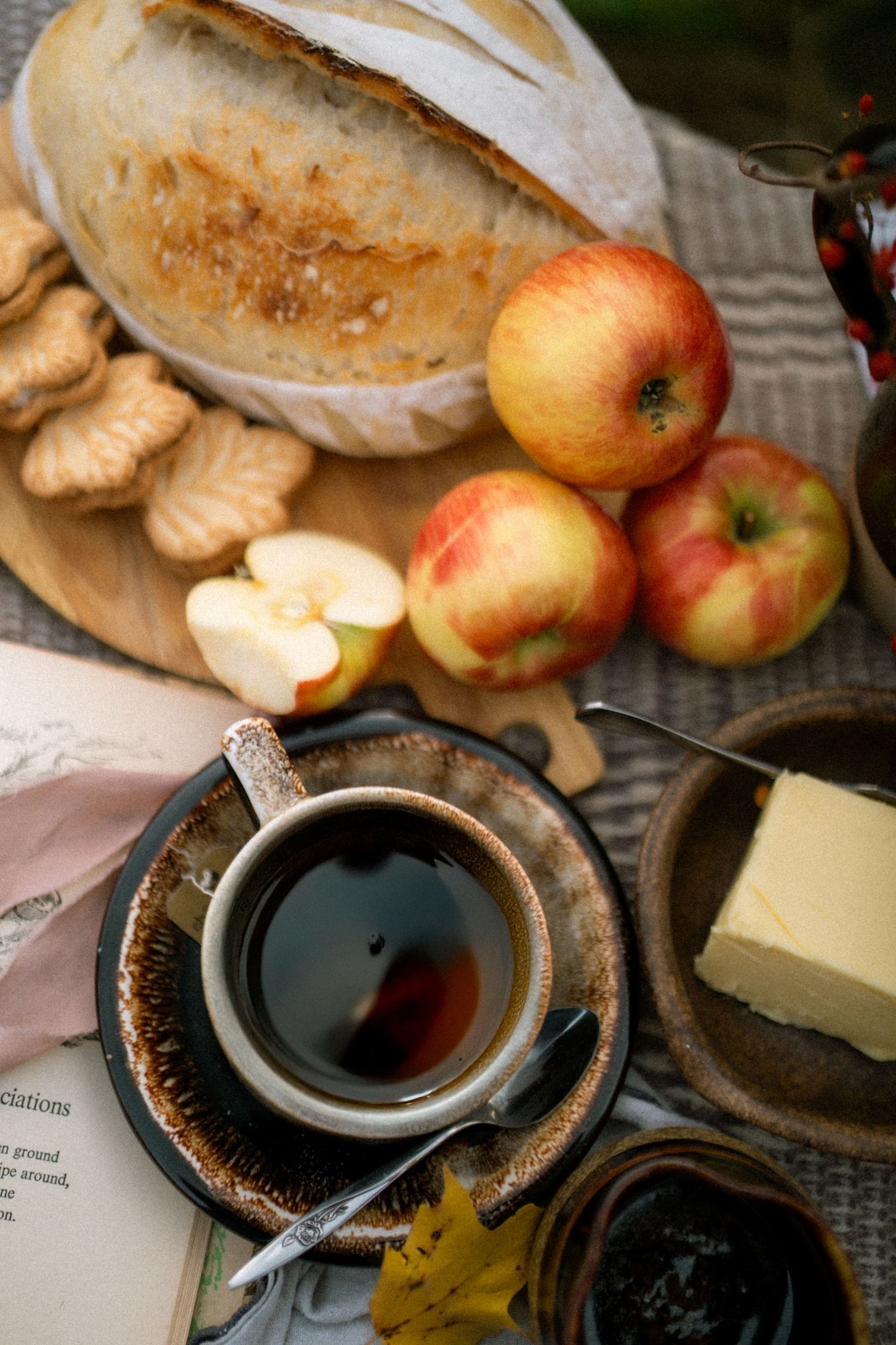 Rustic breakfast scene with fresh bread, apples, butter, and black coffee on a weathered wooden table.