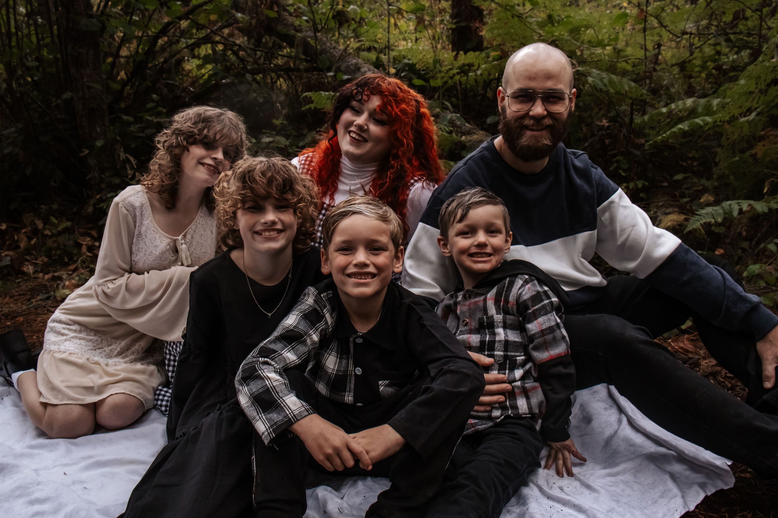 Family of six smiling together on a blanket in a lush green forest setting.