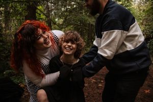 Woman with red hair and man playfully tickling a laughing child in the forest.