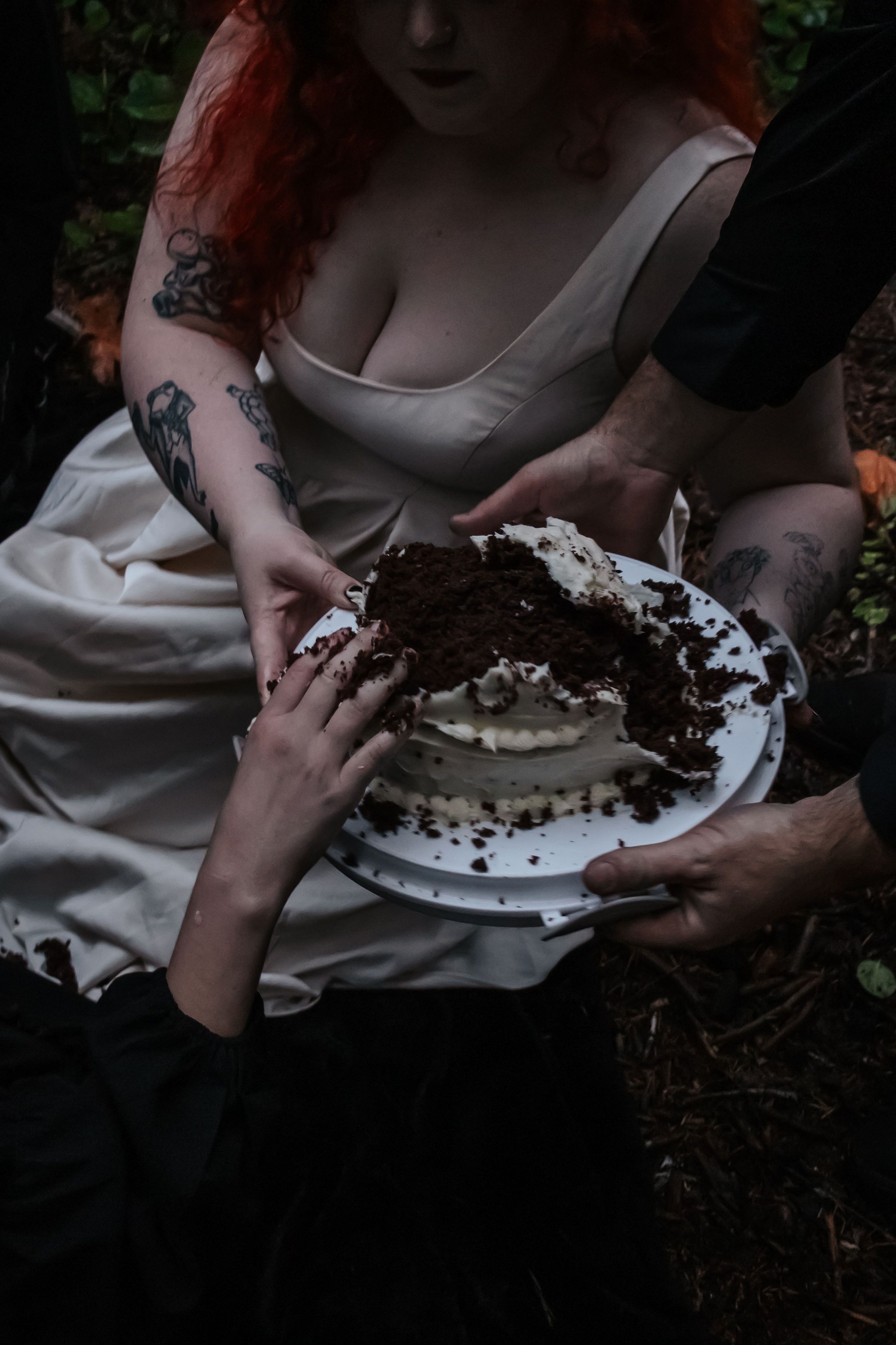 Tattooed woman in white dress holding a chocolate and cream cake in a forest.