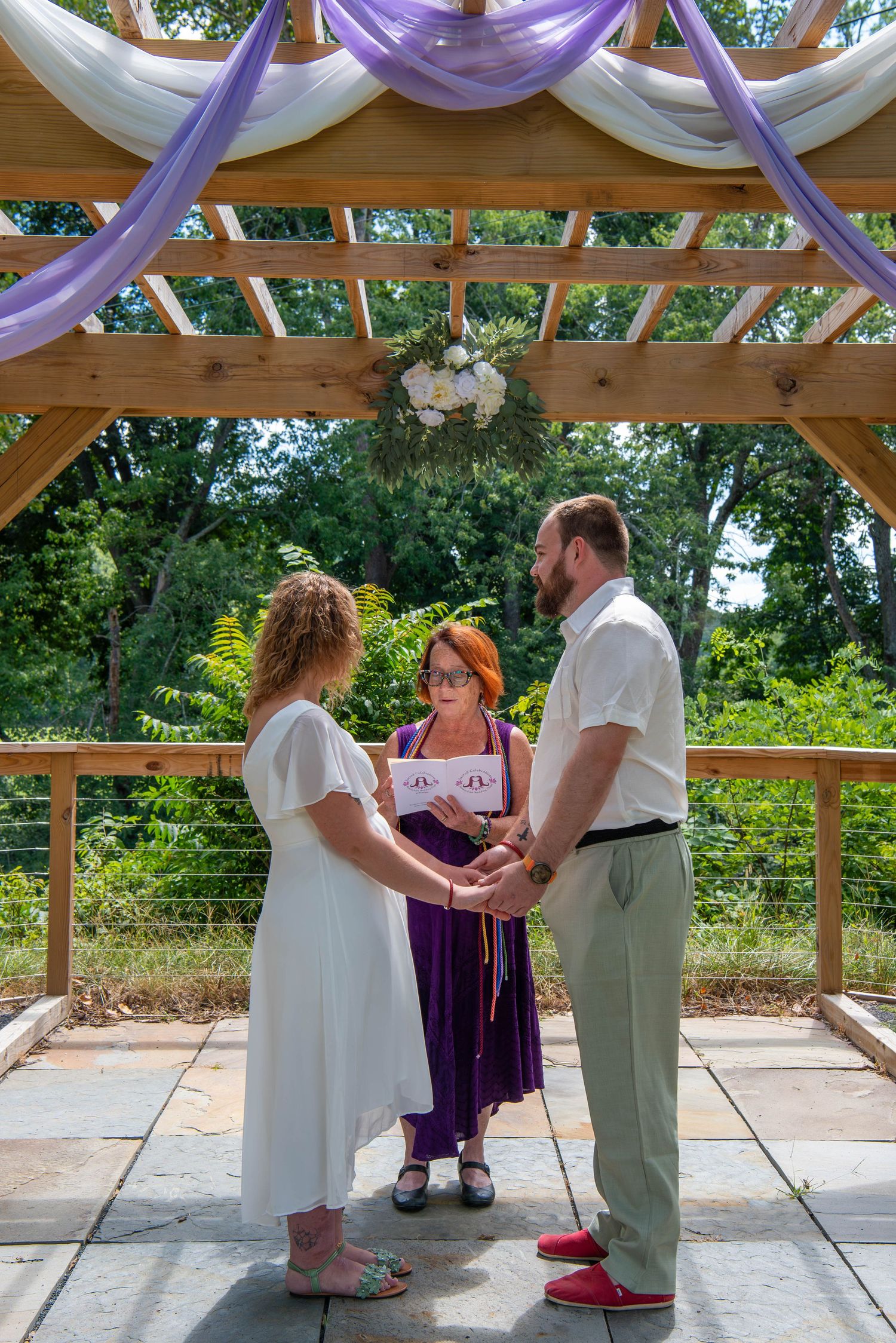 A wedding ceremony takes place under a wooden pergola decorated with purple and white fabric draping.