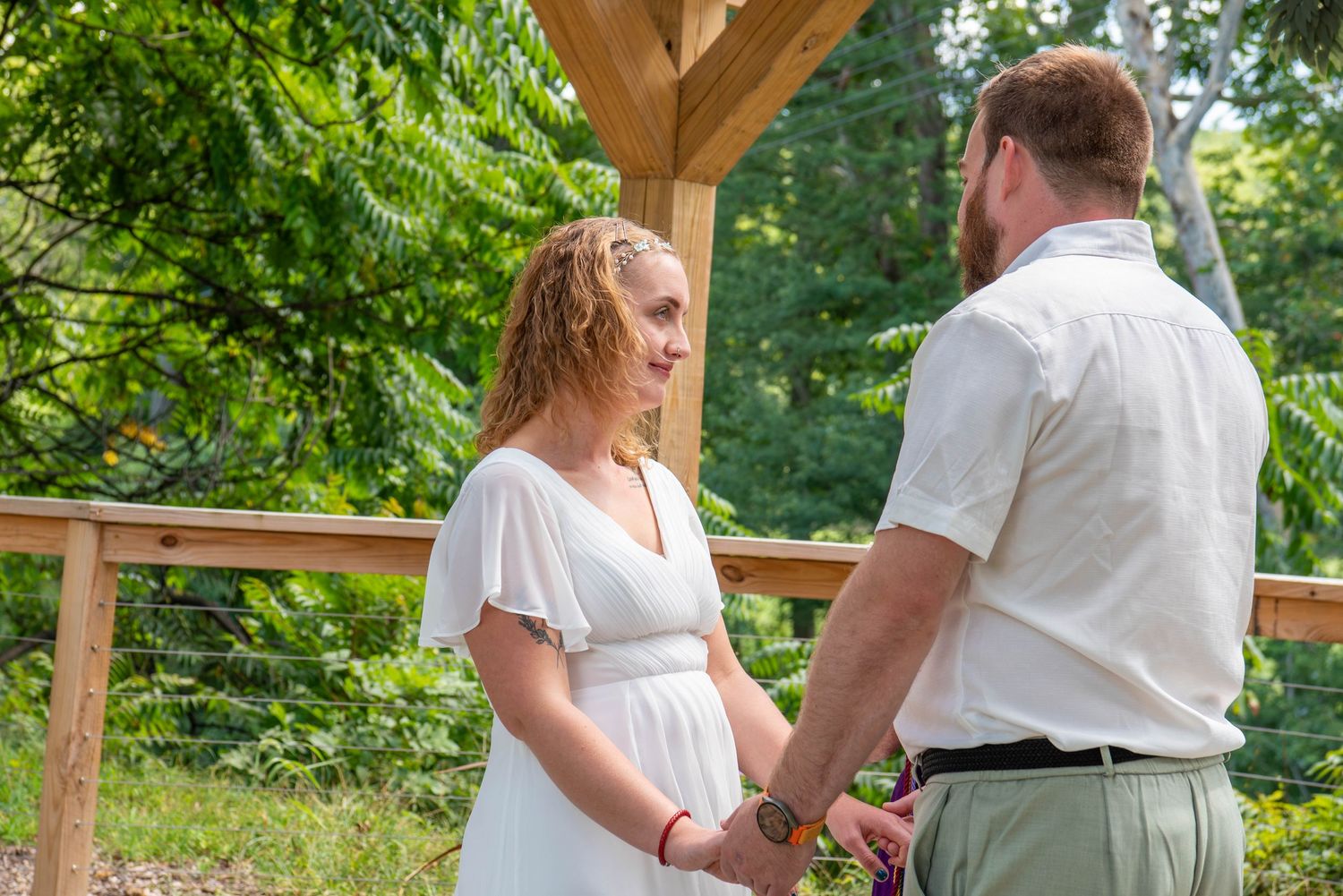 A couple stands together at an outdoor wedding ceremony under a wooden pergola surrounded by greenery.