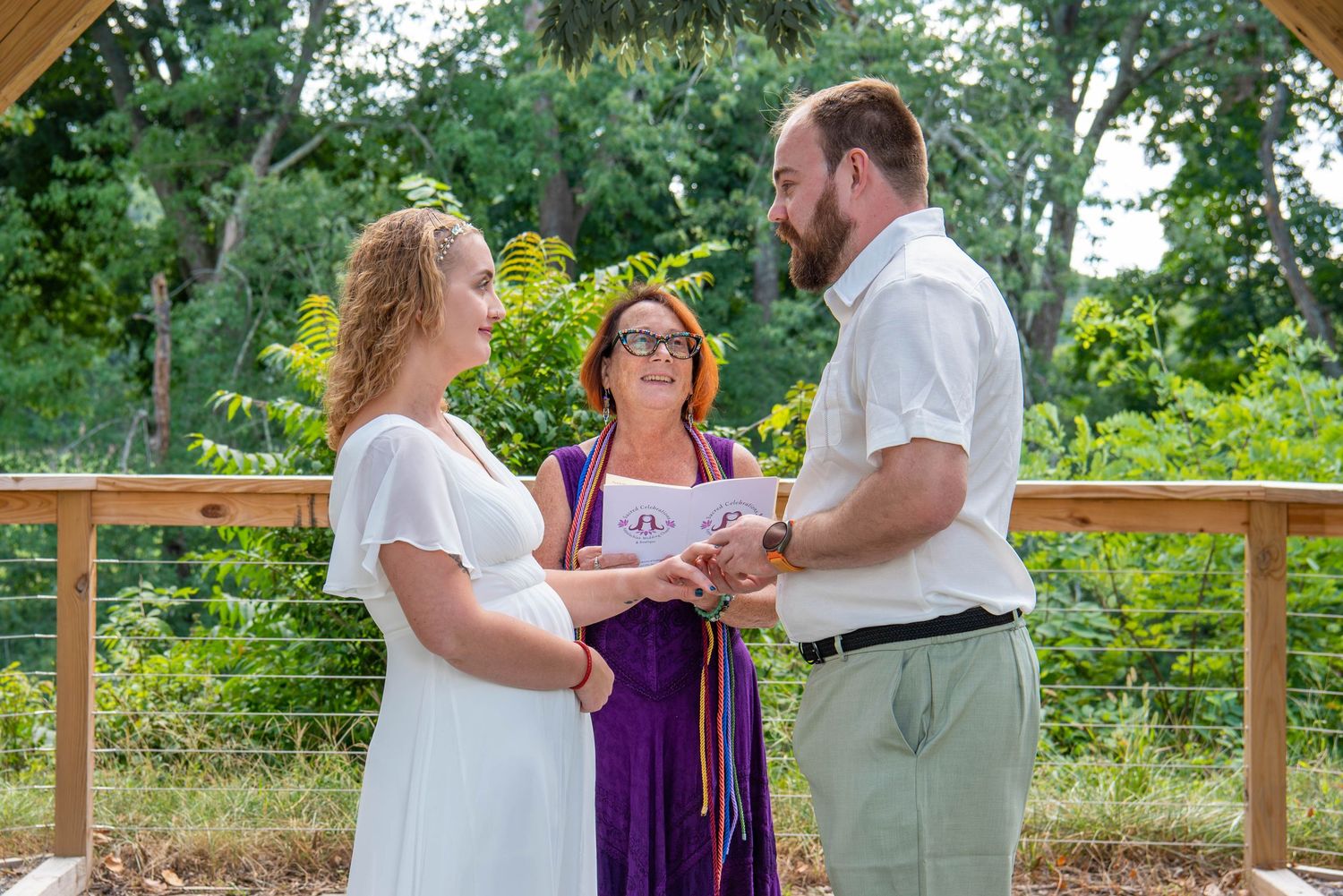 A couple exchanges rings during an outdoor wedding ceremony with an officiant in purple on a wooden deck.