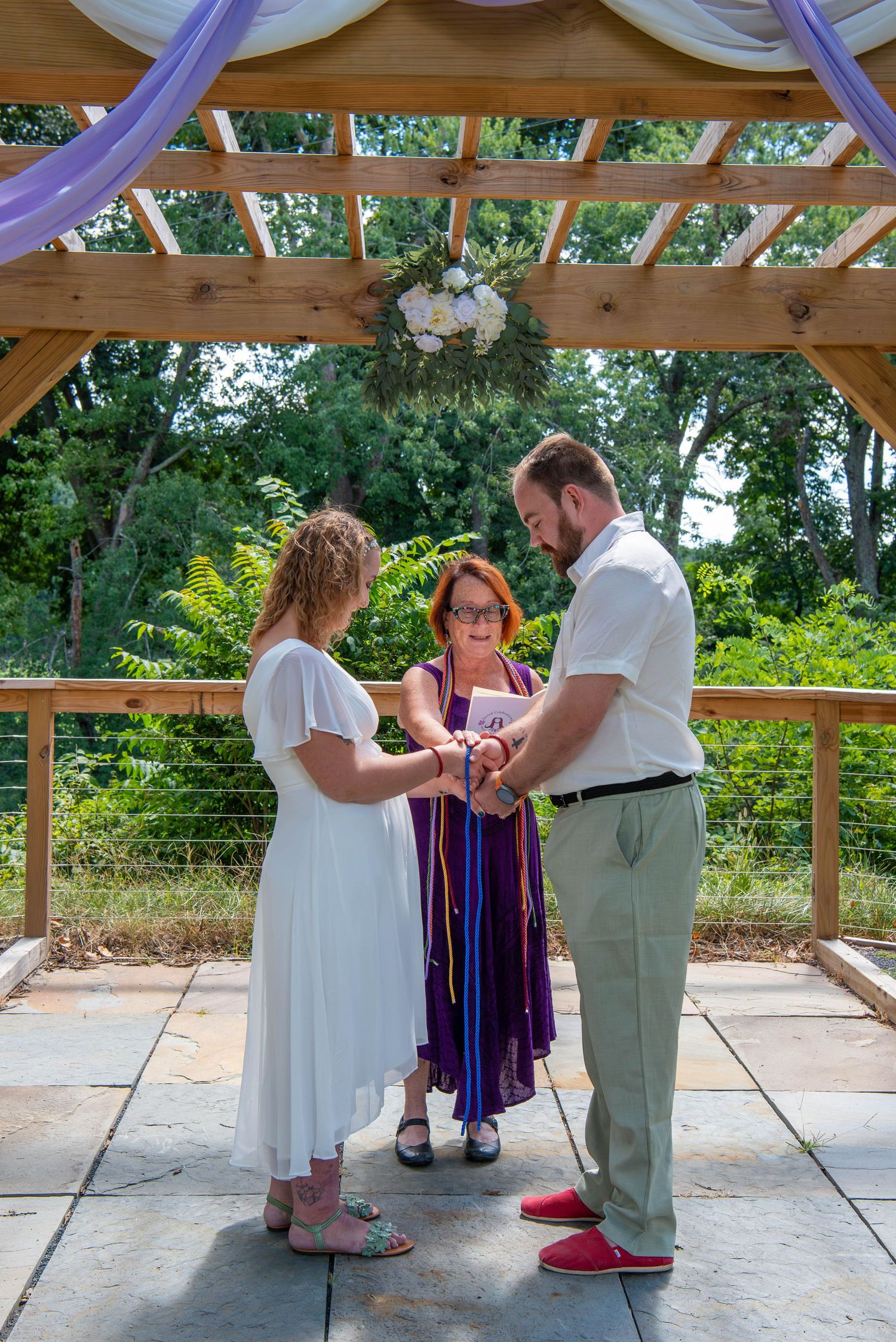 A wedding ceremony taking place under a wooden pergola with purple and white decorative elements in an outdoor setting.