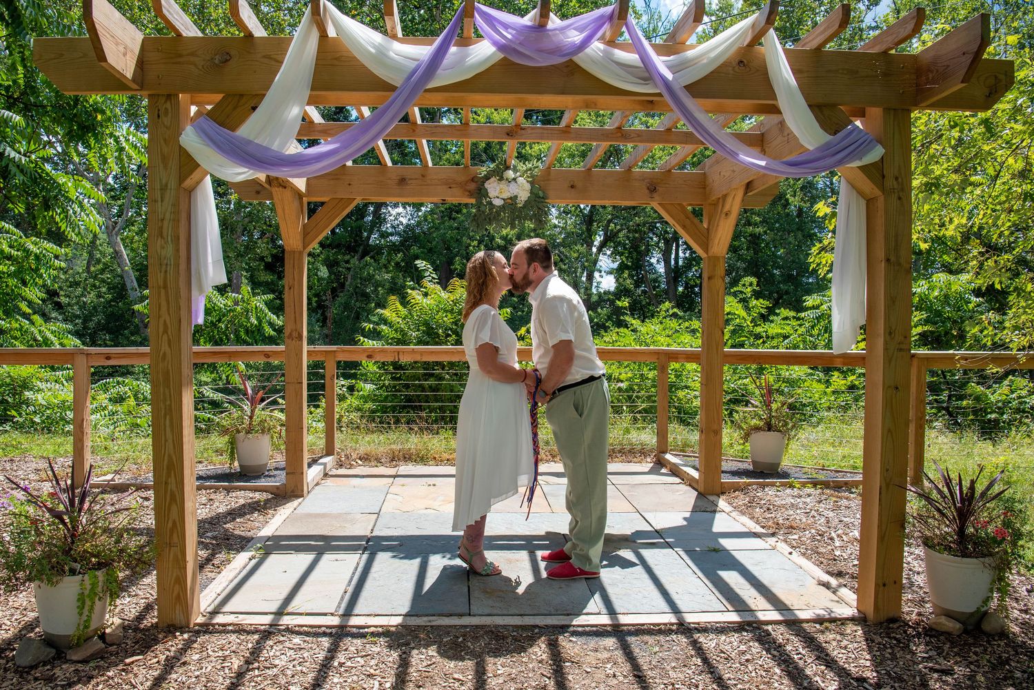 A wooden pergola structure stands in a lush garden setting with dappled sunlight filtering through.