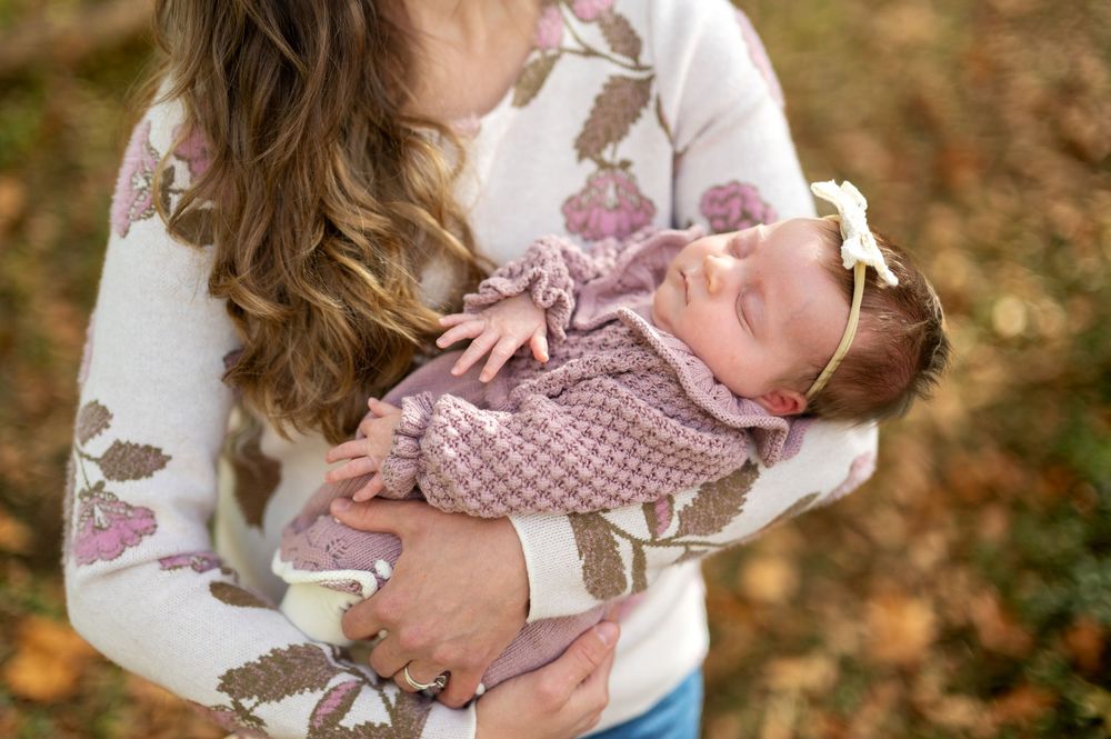 Mother holding sleeping newborn baby dressed in pink knit outfit with bow headband outdoors in autumn.