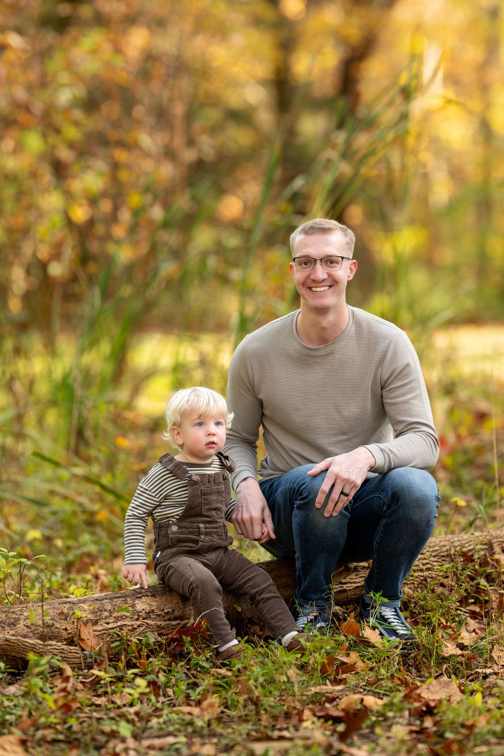 Father with glasses crouching beside toddler son sitting on log in colorful fall forest setting.