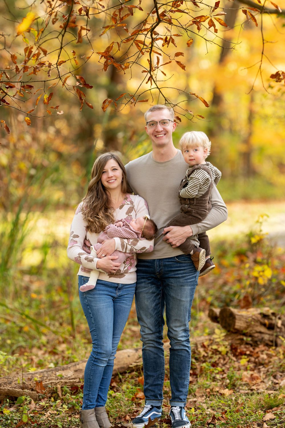Happy young family of four with newborn and toddler posing under fall foliage in golden forest.