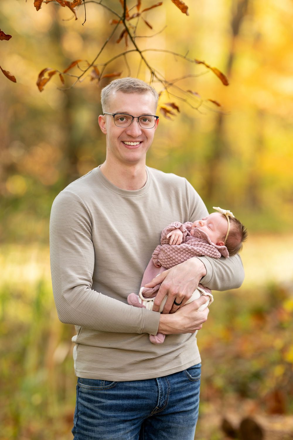 Proud father in glasses smiling while gently cradling sleeping newborn in pink outfit outdoors in fall.