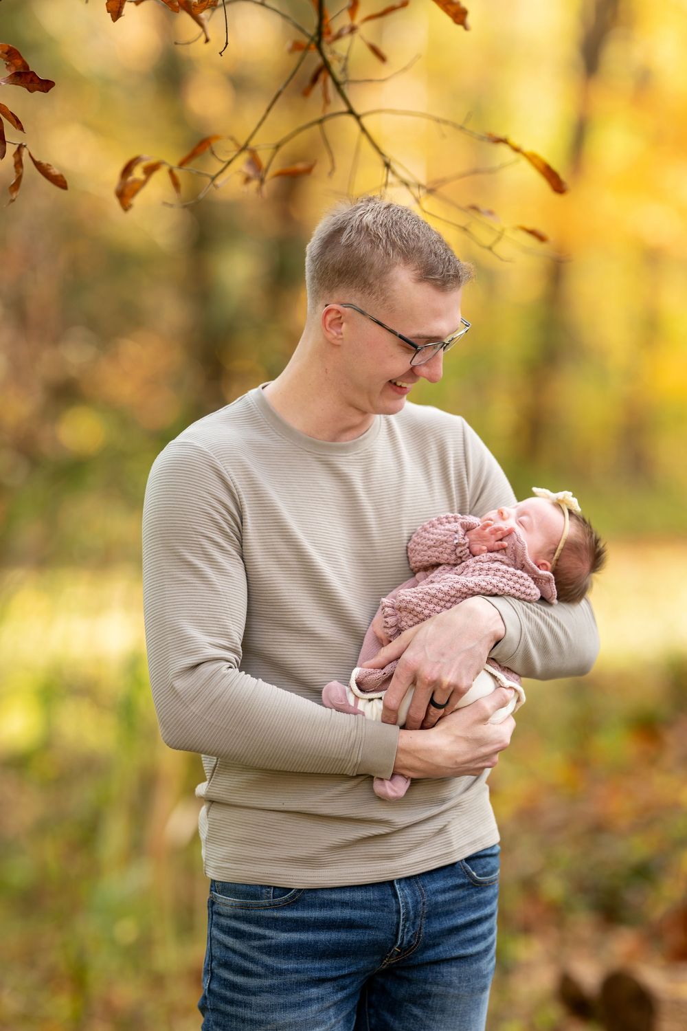 Father in beige sweater lovingly gazing down at sleeping newborn baby in autumn park setting.