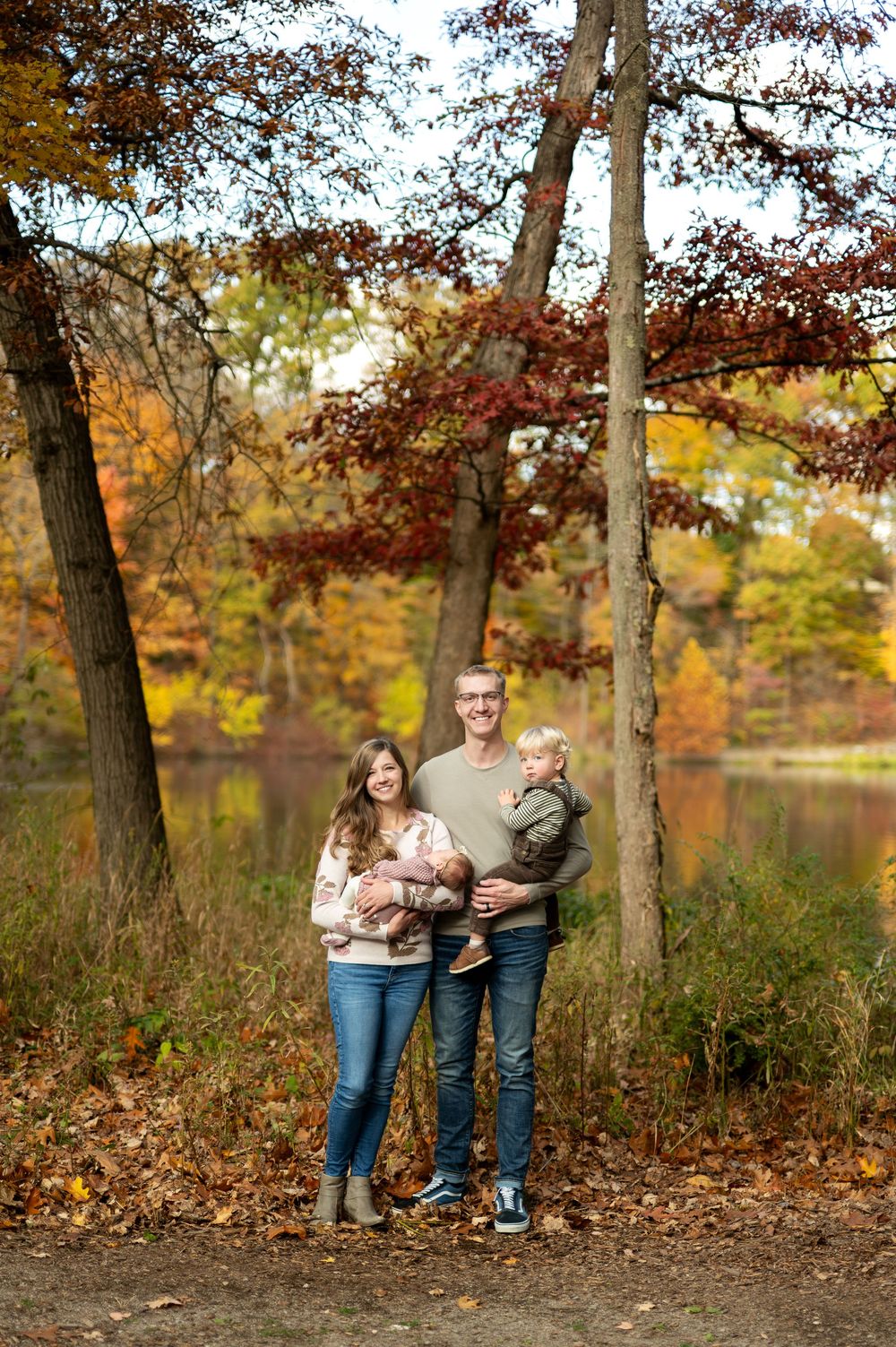 Young family with newborn baby and toddler smiling together in a vibrant autumn woodland park.