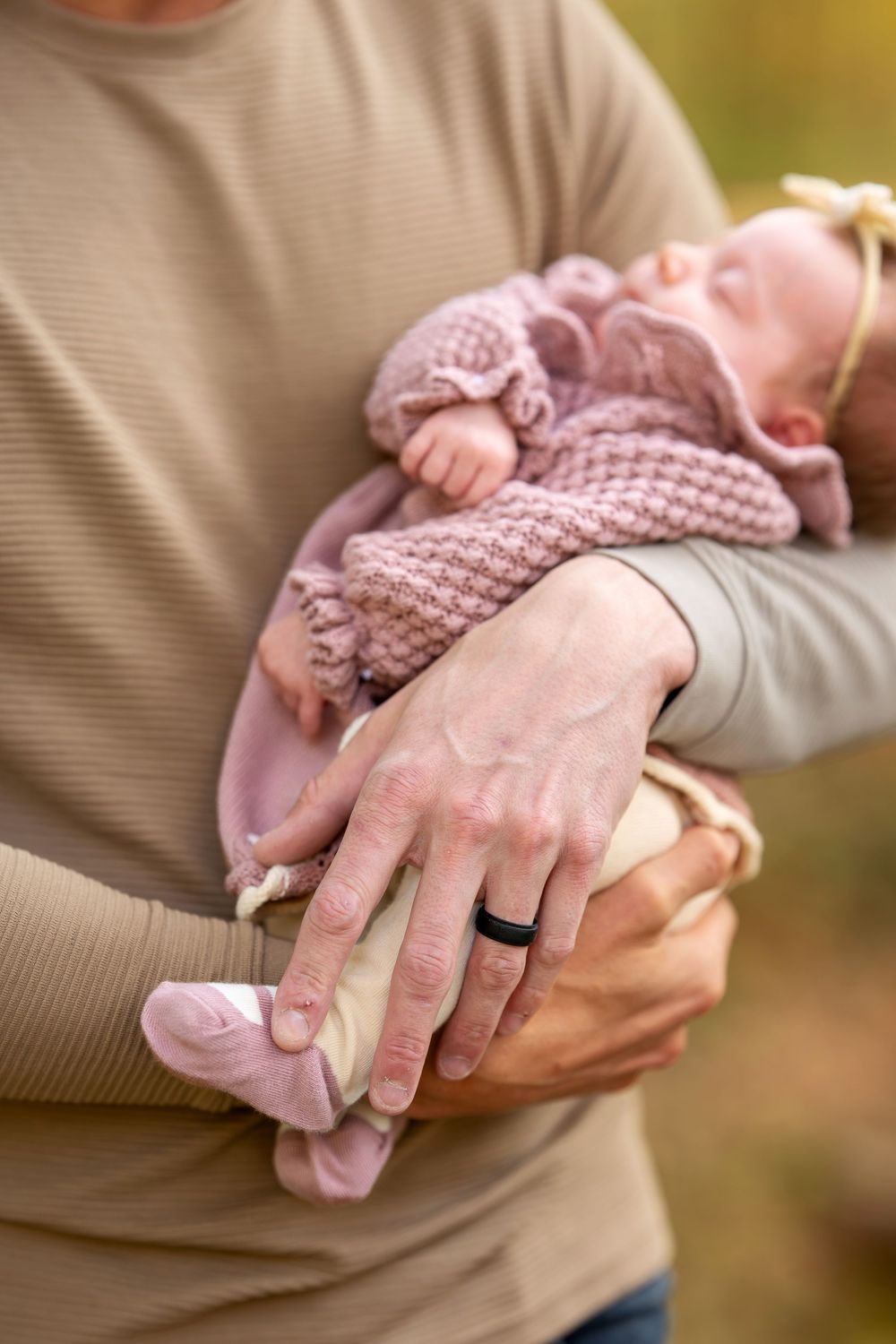 Close-up of parents hands cradling sleeping newborn in pink knit outfit with father wearing black ring.