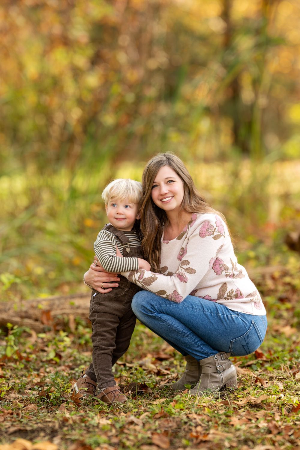 Smiling mother crouching and hugging blonde toddler son in autumn park with colorful fall foliage.