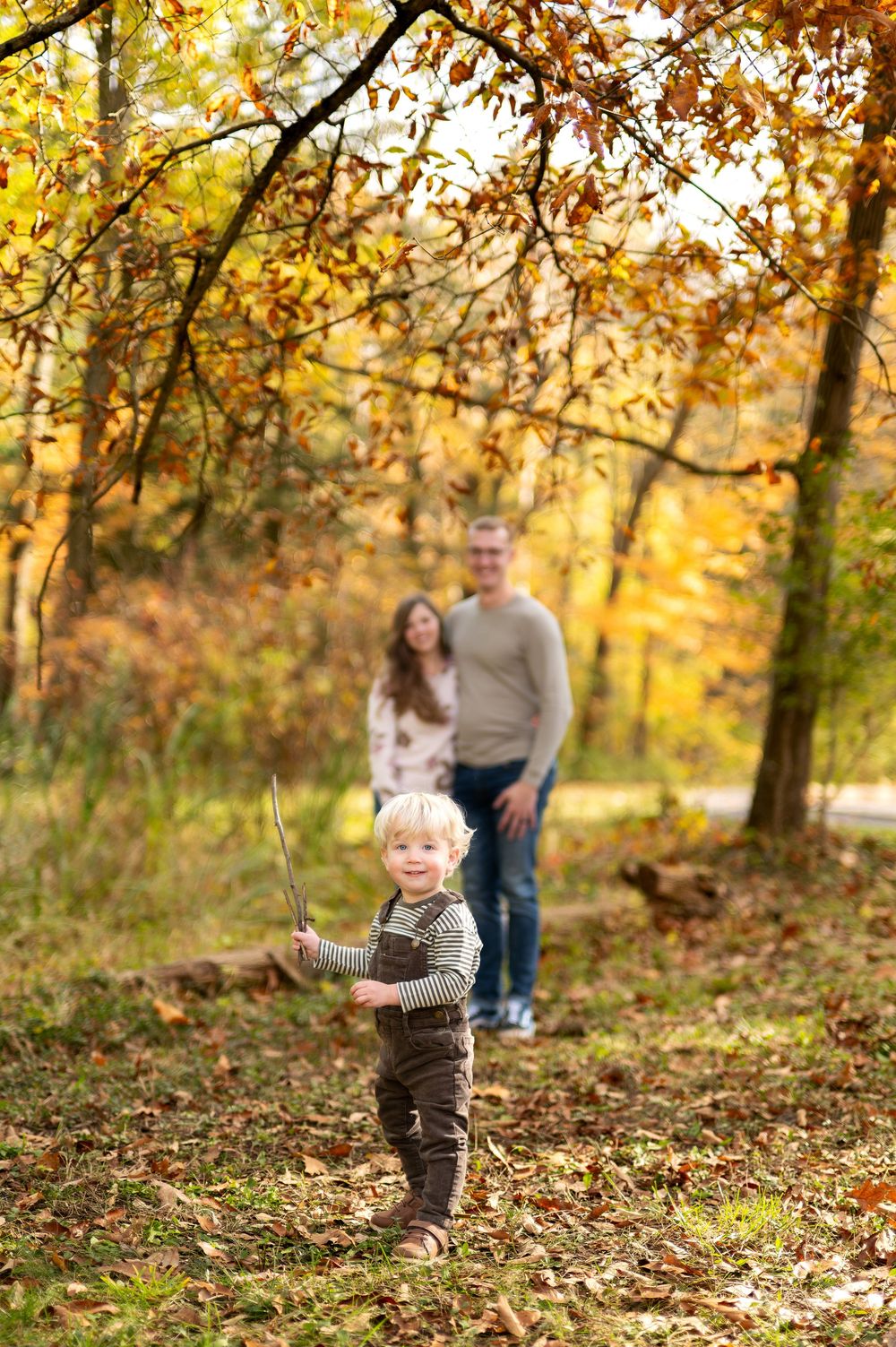A happy toddler holds a stick while his parents watch in a colorful fall forest.