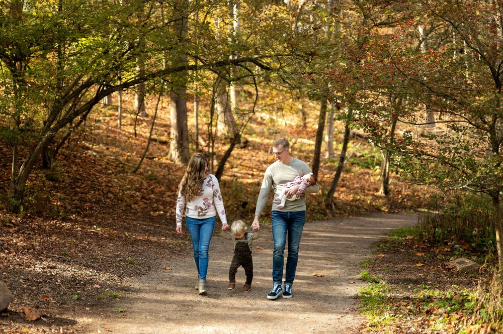 A family walks hand-in-hand with their toddler and dog on a fall forest trail.