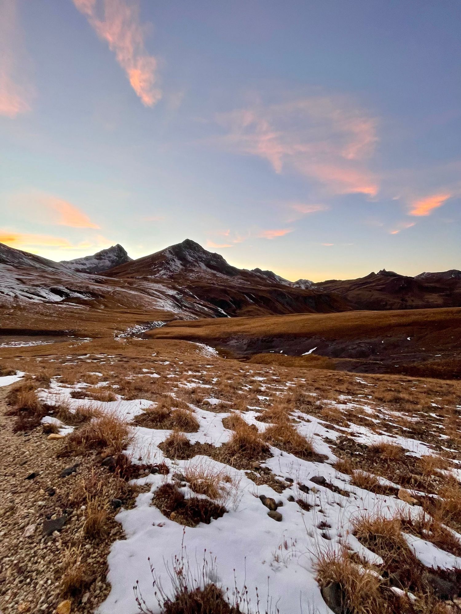 Panoramic view of snow-dusted mountain peaks and rolling hills at sunset with pink clouds in the sky.