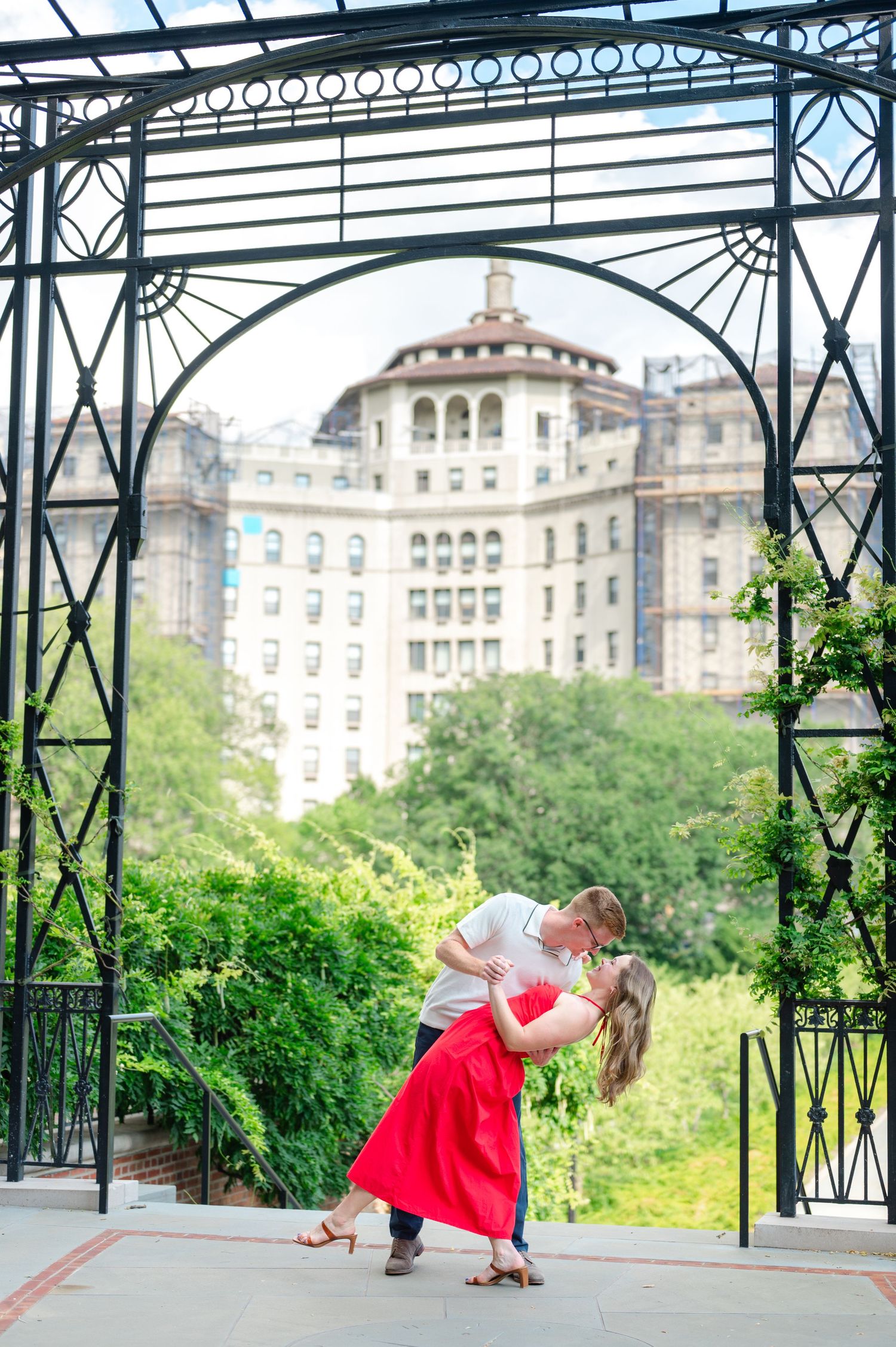 A couple dances under a decorative metal gazebo structure with historic buildings visible in the background.