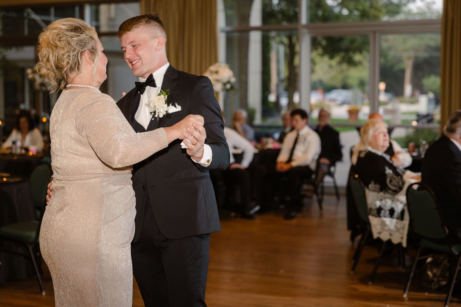 A couple shares a dance on a wooden floor at an elegant reception venue with other guests seated in the background.