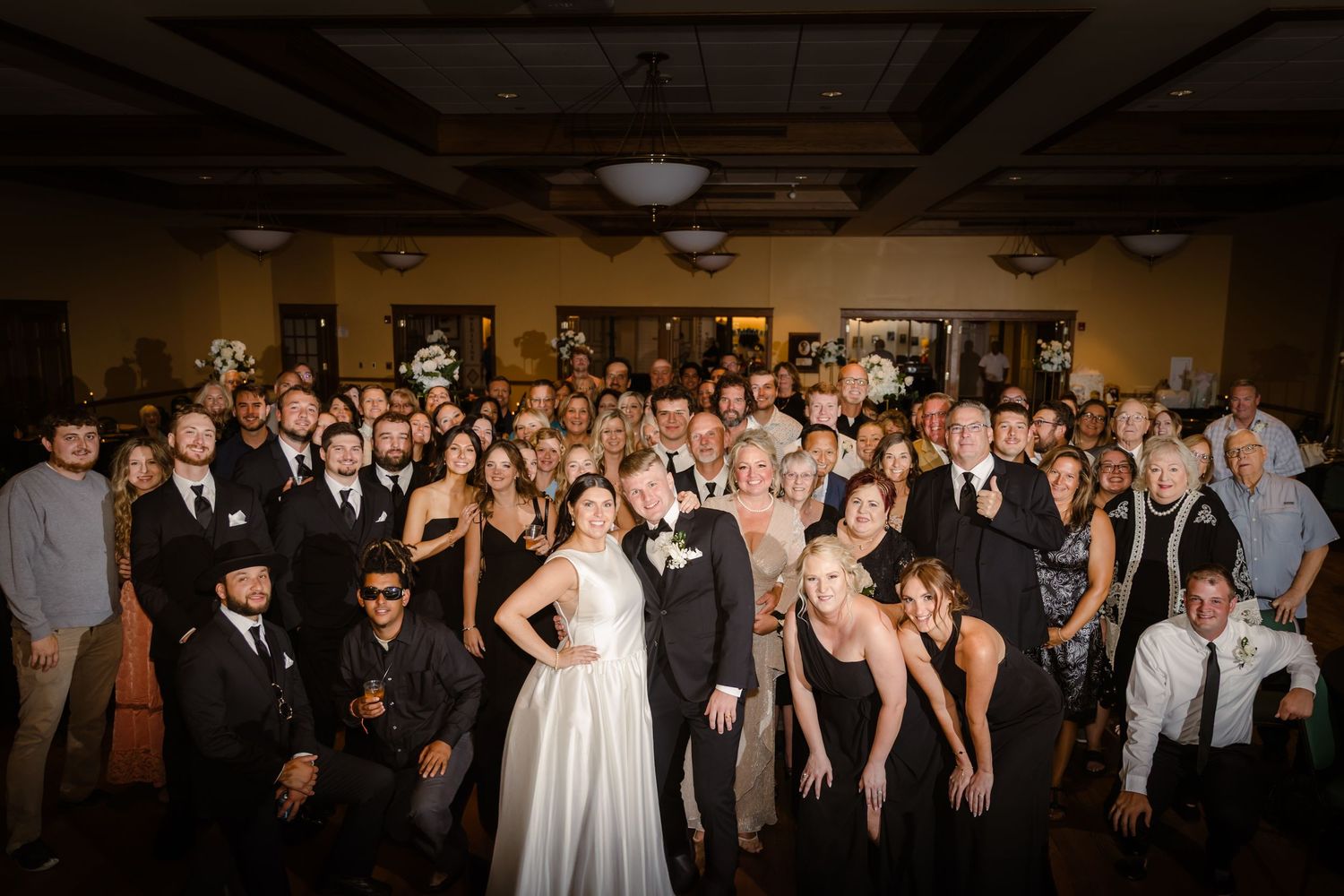 Large wedding party group photo with guests and wedding couple gathered together in a dimly lit reception venue.
