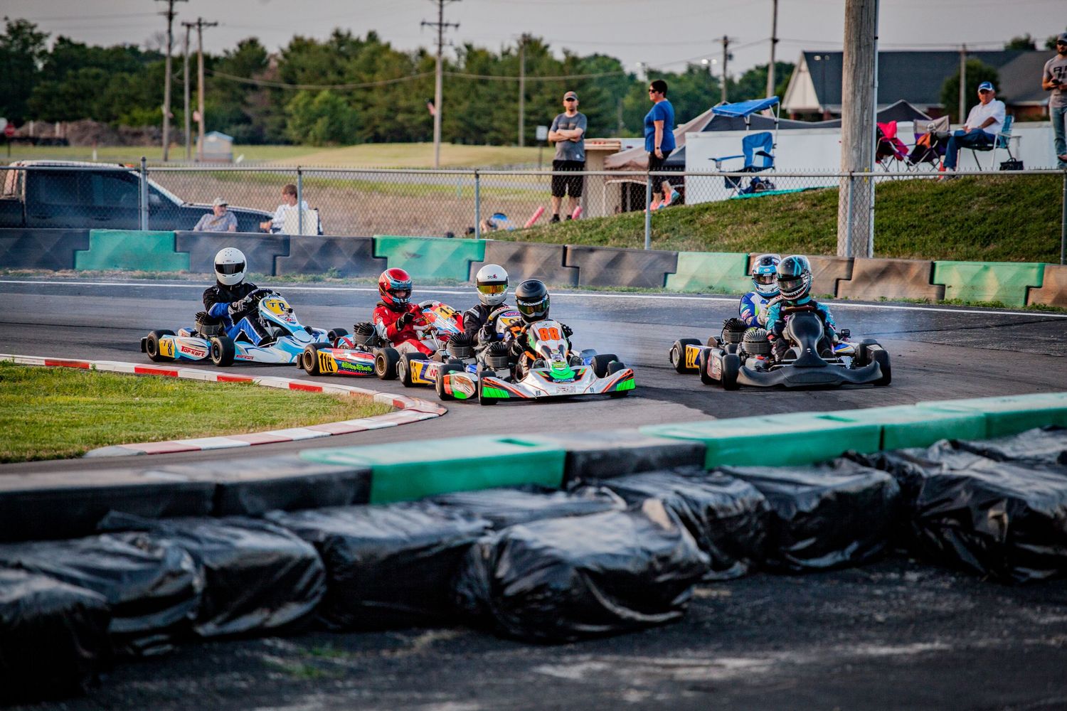 Go-kart racers navigate a turn on an outdoor track with tire barriers and safety barriers visible during a competition.