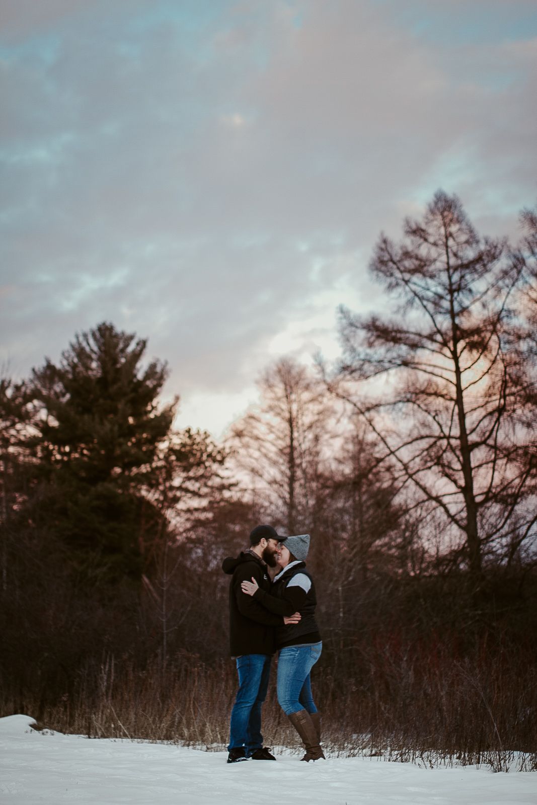 A panoramic winter scene shows two people in dark winter coats walking through a snowy landscape at dusk.
