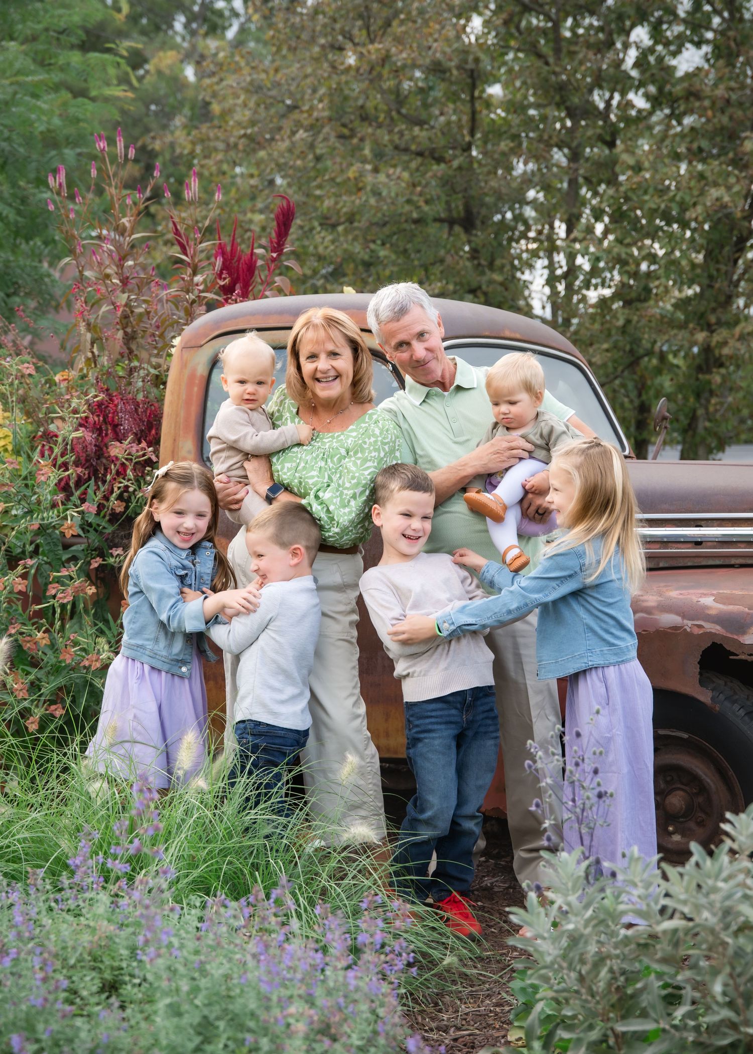 Family laughs and plays together in a garden with purple flowers and a vintage truck in the background.