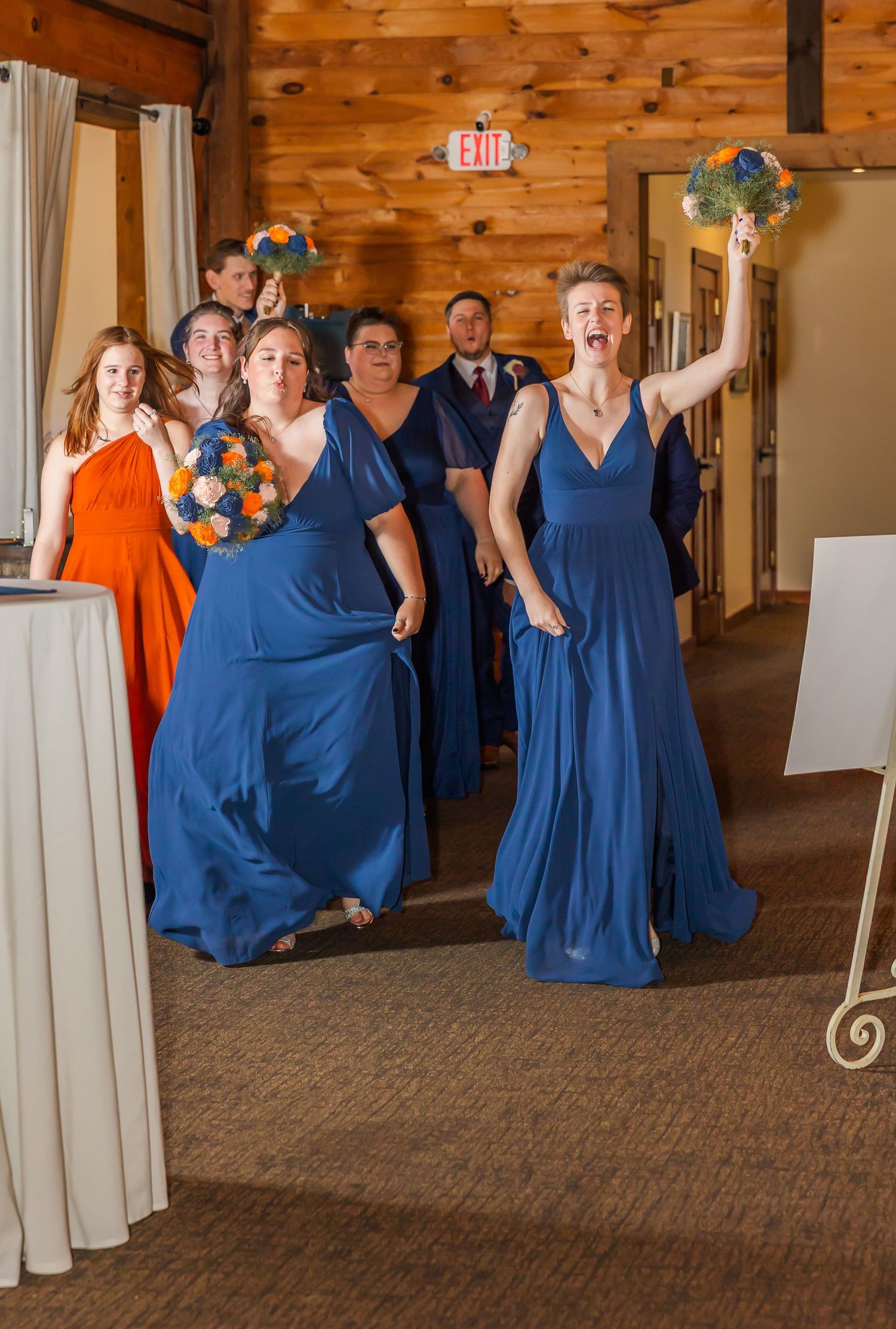 Wedding party members in navy blue dresses dance and celebrate in a rustic wood-paneled venue.