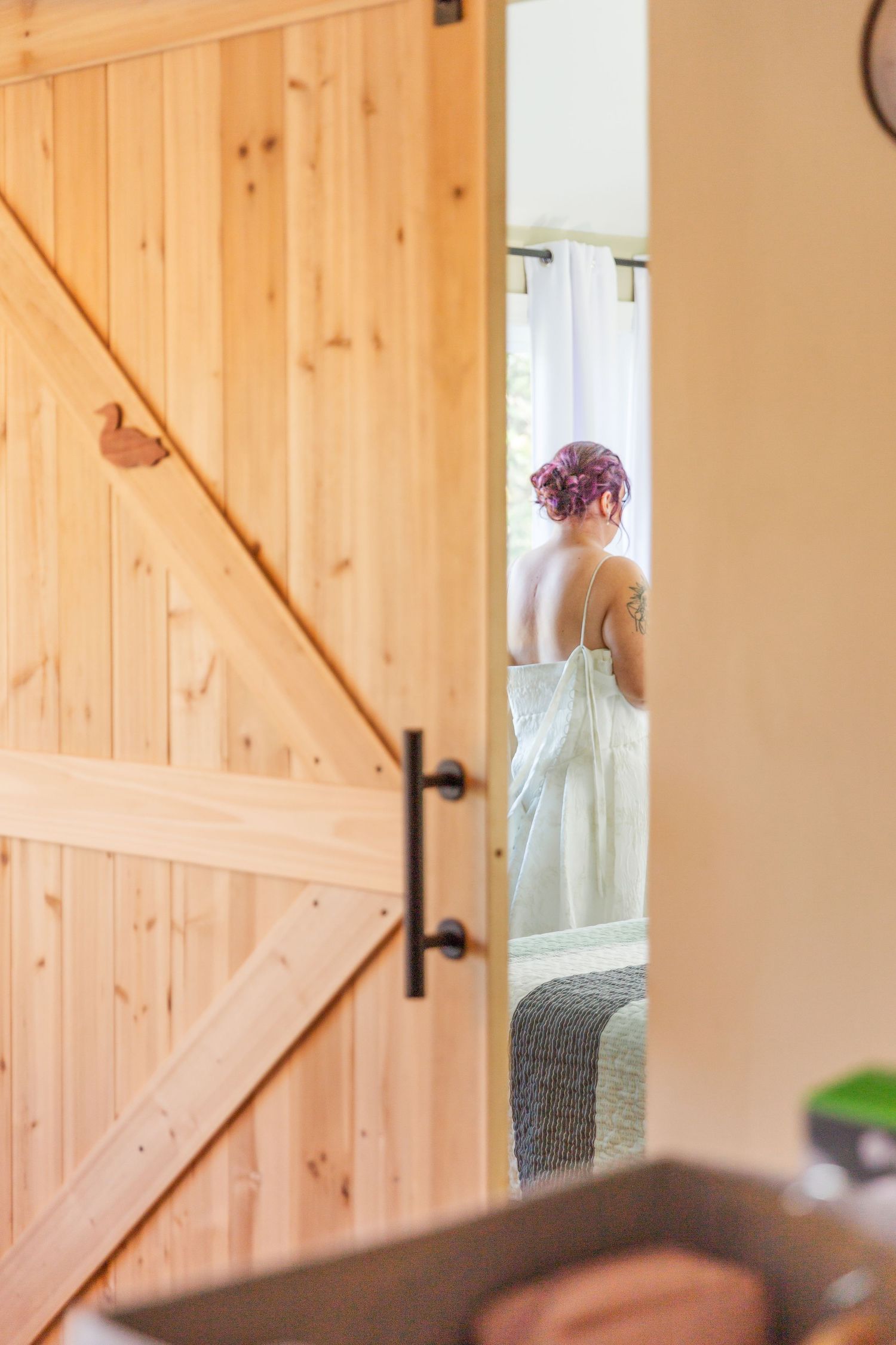 A glimpse through a wooden barn door shows someone in white with pink hair standing in a sunlit room.