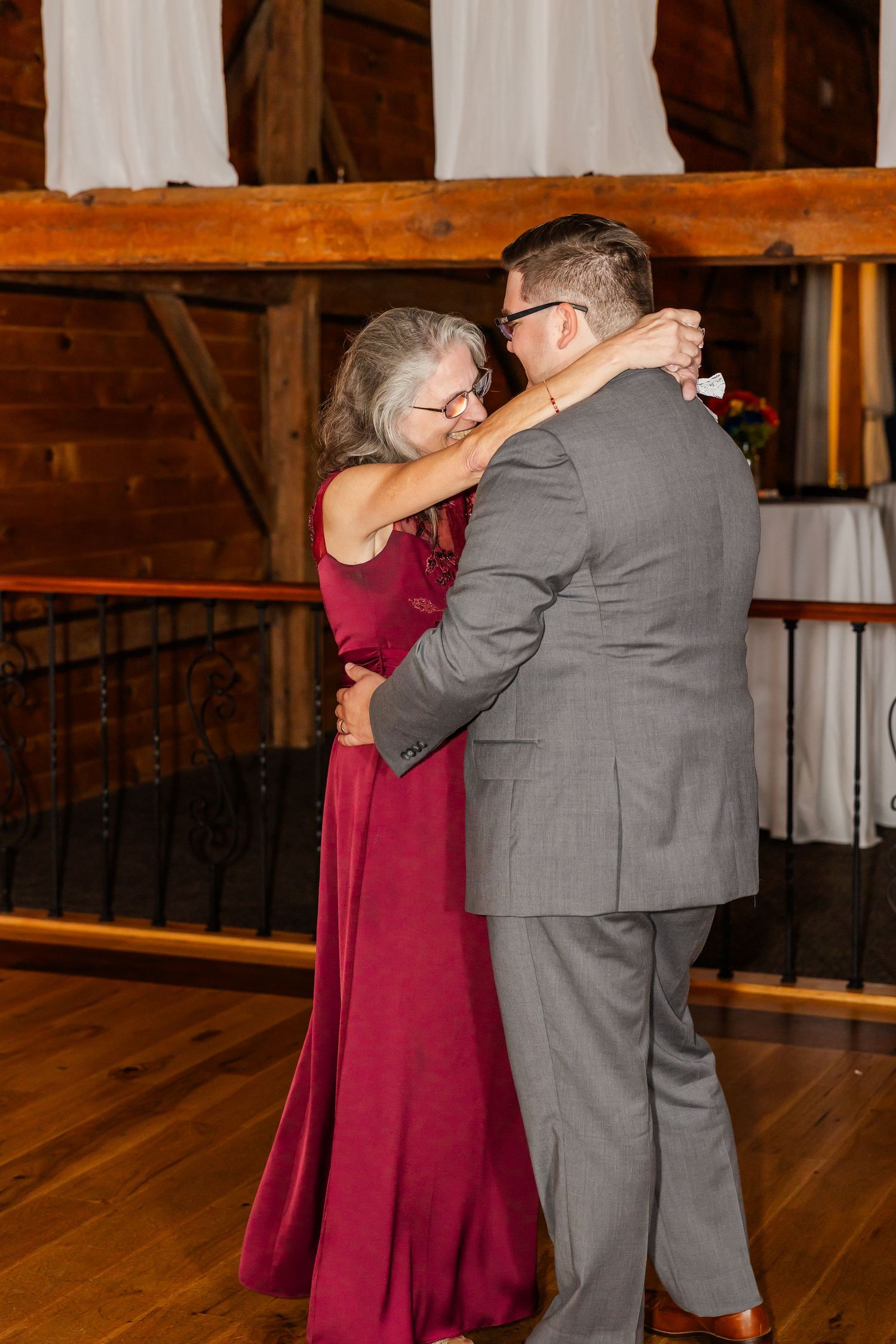 A couple in burgundy dress and gray suit share an intimate dance moment in a rustic wooden venue.