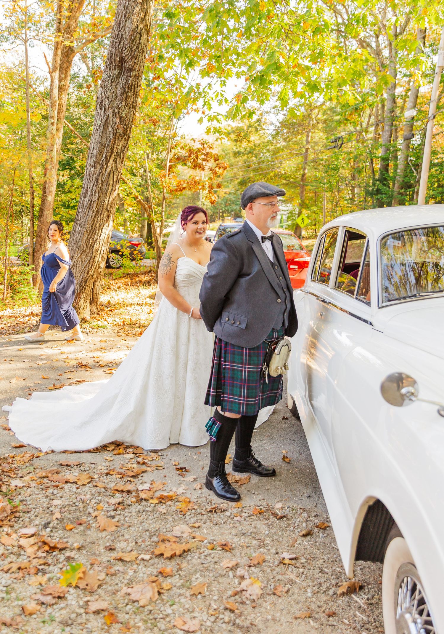 A wedding party gathers around a classic white vintage car on a leaf-covered autumn forest path.