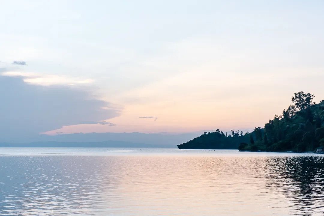 Peaceful lake at sunset with pastel sky and tree-covered shoreline in the distance.