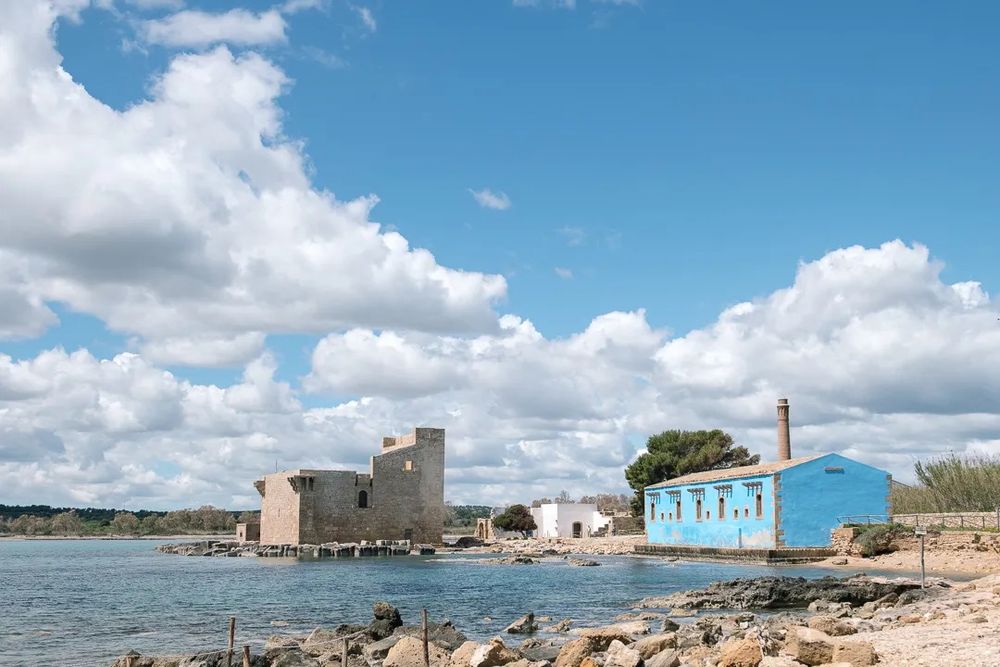 A historic stone tower and a small blue waterfront building beside the sea under scattered clouds.